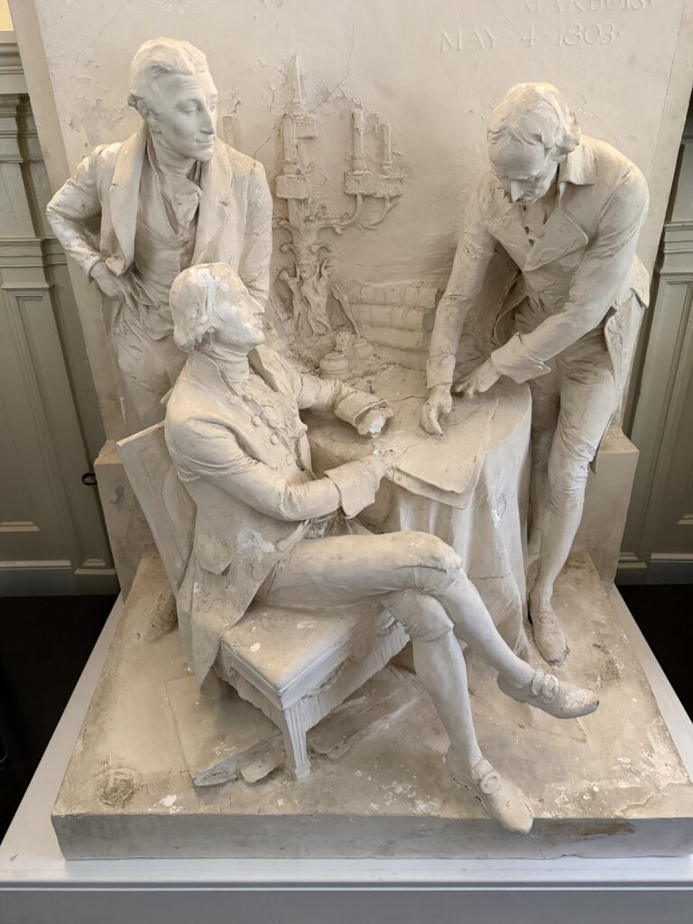 A plaster mold of a sculpture featuring one man sitting down at a table, and two standing up next to him.