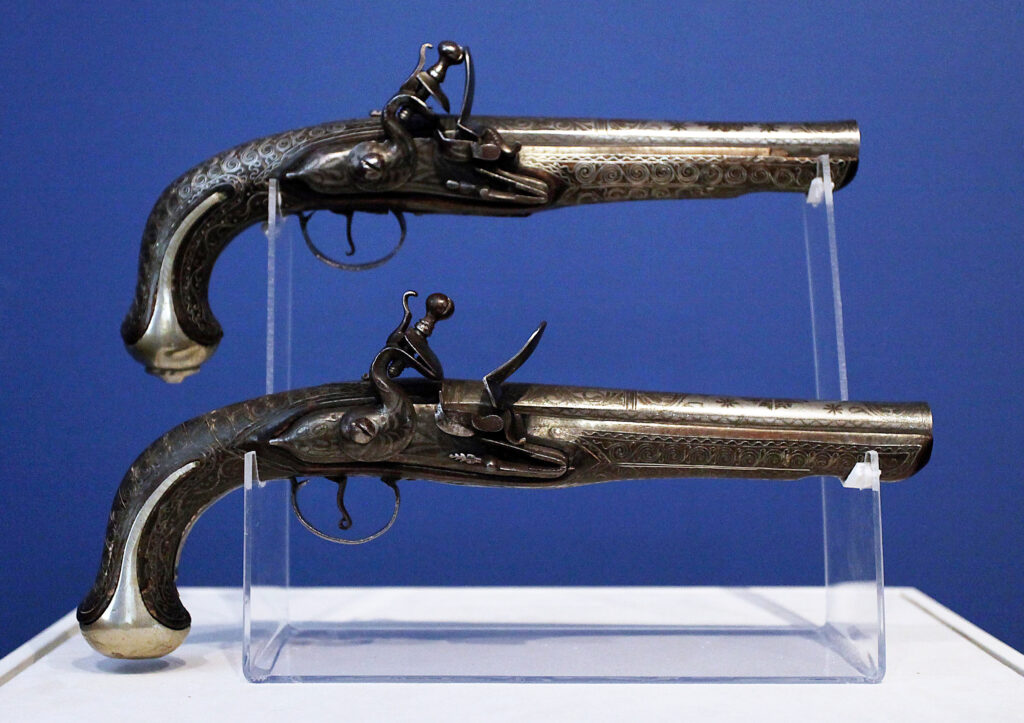 A pair of ornate flintlock pistols with carvings on the handles displayed on a plastic holder